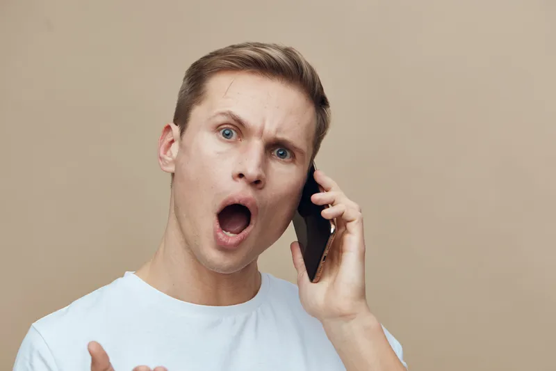 shocked young man talking phone headset surprised emotionally mixed race casual white t-shirt beige background digital detox concept