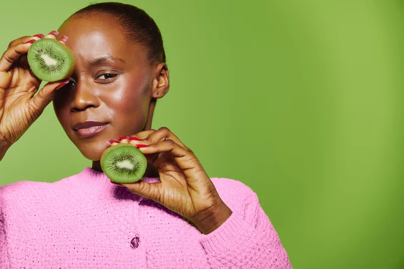 African woman with dark skin holds two halves of fresh kiwi fruit near her face. She wears a pink sweater and poses against a bright green background. Concept of healthy lifestyle and natural beauty.
