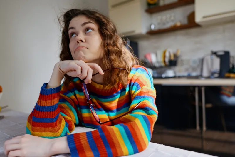 Cheerful woman indoors, relaxed lifestyle at home, colorful sweater, thoughtful expression, sunlit kitchen vibe, casual pose, cozy ambiance, natural colors, everyday moment