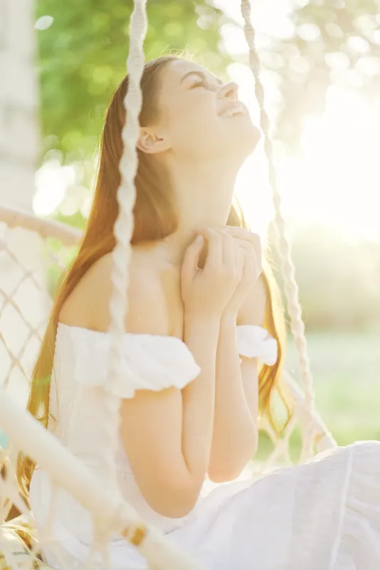 Beautiful young woman in a white dress smiling joyfully in a sunlit outdoor setting, evoking feelings of happiness and serenity