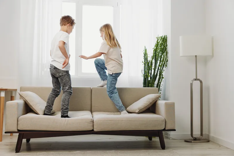 Two children jumping on beige sofa in bright living room with natural light and casual clothing, enjoying playful and energetic moment indoors.