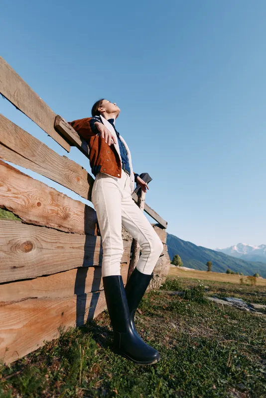 Woman leaning on wooden fence in countryside with mountains and deep blue sky, wearing jacket, scarf and tall boots. Outdoor portrait in meadow, standing pose, sunlight and relaxed leisure.