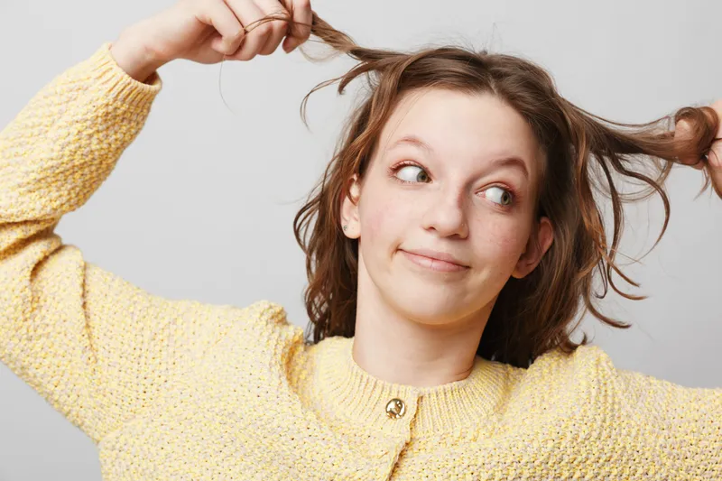 Young woman in yellow sweater pulls her hair playfully, looking to the side with a curious expression. Studio portrait on light background. Concept of fun, youth, and casual style.