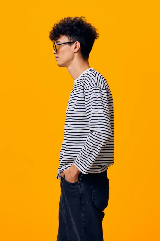 Young man in striped shirt standing in profile with hands in pockets against bright orange background, wearing glasses and black jeans, casual style.