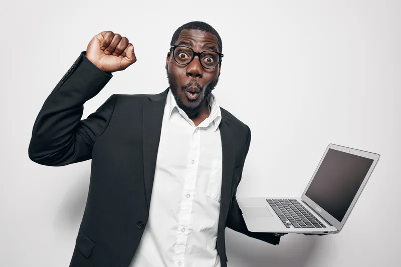 African American man with dark skin wearing glasses and black suit holding laptop and celebrating success with raised fist and surprised face on white background.