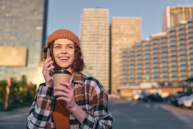 Woman with coffee and phone in city smiling during golden hour, candid lifestyle portrait with beanie and plaid jacket, authenticity and mindful living, emotional storytelling mood.