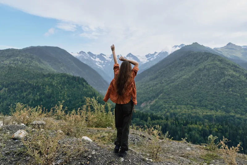 Woman hiker stands on a rocky overlook with arms raised, enjoying vast mountains and landscape, showcasing outdoor nature, freedom, and triumph in pristine scenery