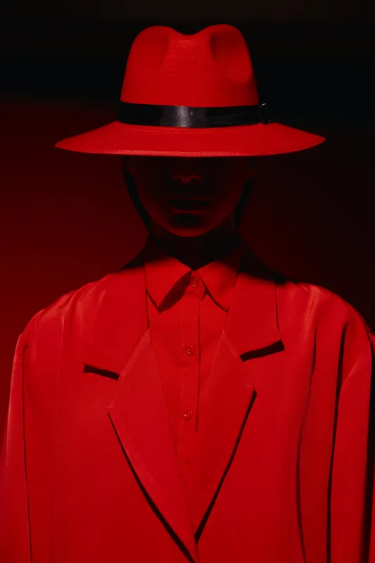 Woman in stylish crimson hat and coat faces camera under dramatic red lighting with shadowed facial features. Creative fashion portrait concept with strong use of color contrast and mood.
