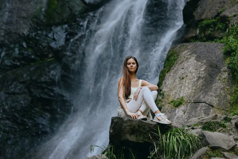 Young woman in white activewear sitting on rock near waterfall surrounded by green plants and moss, enjoying peaceful nature moments outdoors in summer.