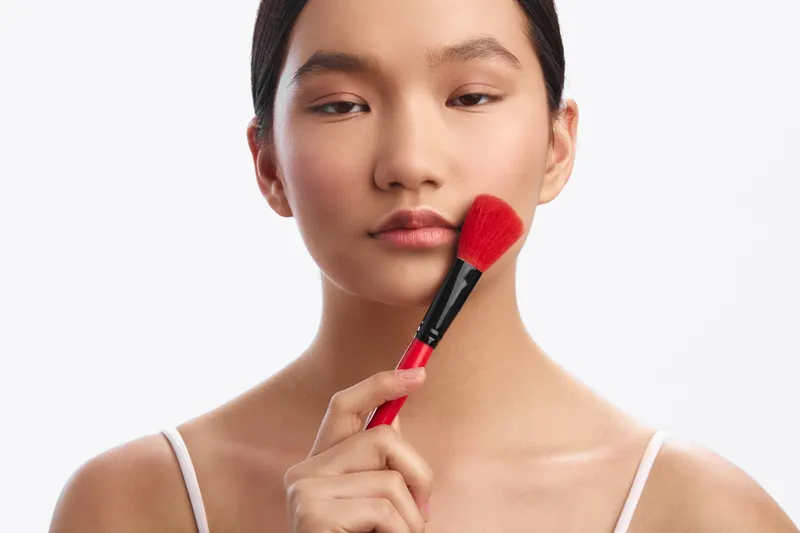 Asian woman applies makeup with a soft brush in a clean studio setting, presenting a calm beauty focus and thoughtful concentration for an intimate portrait.