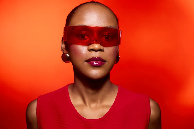 Elegant African woman with dark skin wearing red sunglasses and sleeveless top poses confidently against bright red background. Concept for fashion, style, and modern beauty.