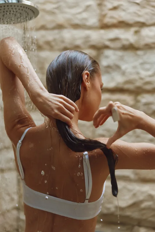 woman showering outdoors with wet hair under running water, natural stone wall background, relaxing moment of personal care and freshness