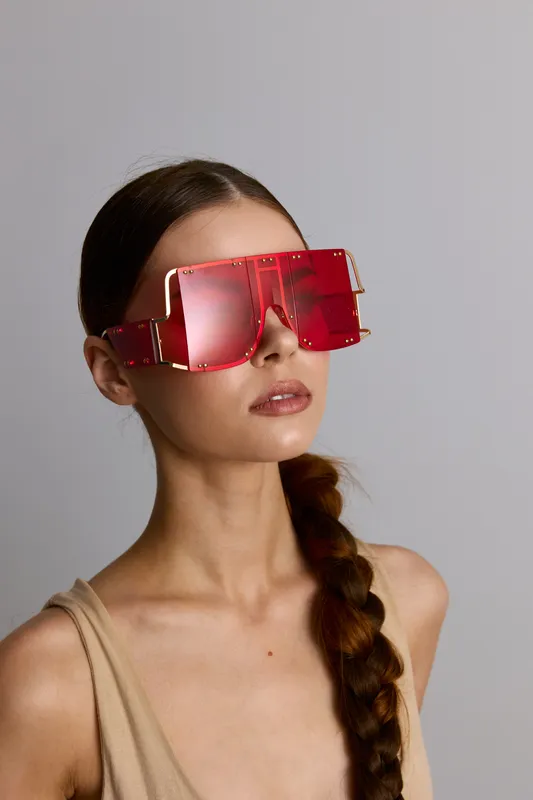 Young woman wearing oversized red futuristic sunglasses with braided hair and beige top against gray background, modern fashion accessory concept.