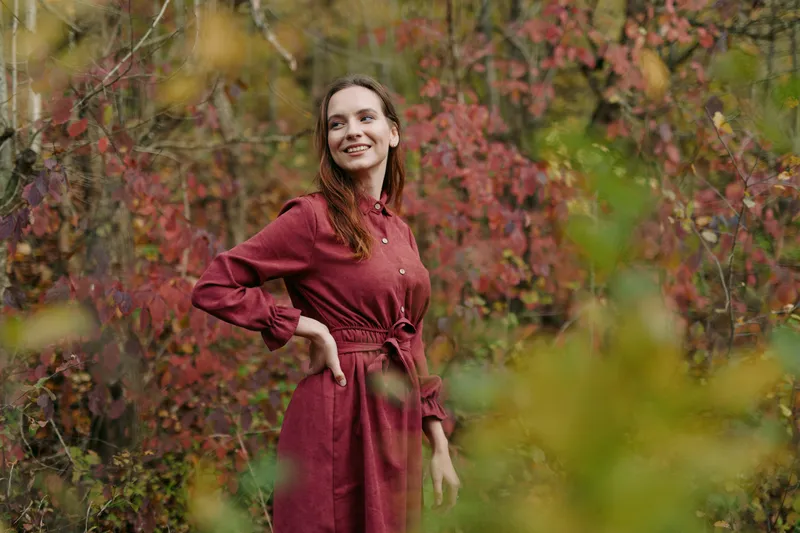 A candid portrait of a smiling woman in a red dress among autumn foliage, conveying authenticity and credibility with genuine expression, natural posture and believable everyday style.