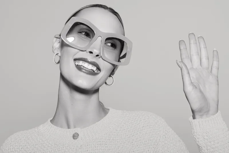 Smiling white woman with pale skin wearing large glasses waves hand in bright studio, happy and confident pose.