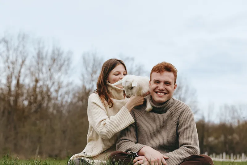 A couple sits on the grass outdoors, sharing a moment with a small goat kid between them. They wear cozy sweaters in a calm countryside setting, conveying tenderness, connection, and simple joy.