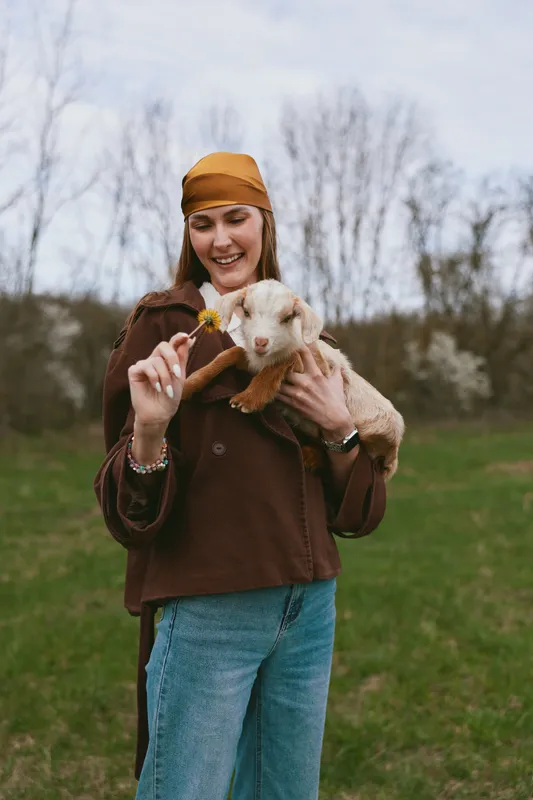 Joyful woman outdoors, feeding a goat kid with a small flower, warm smile, casual sweater, meadow backdrop, natural colors, bright atmosphere, wholesome lifestyle moment