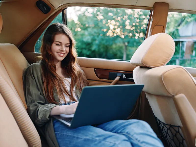 Young woman working on a laptop in a cozy car interior, smiling and enjoying the moment, with warm natural light filtering through the window.