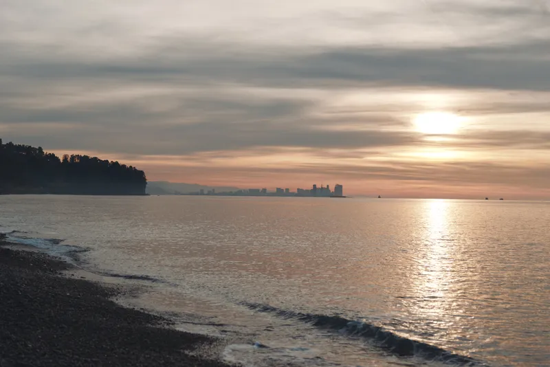 Sunset over calm sea and gentle coastline with a sandy beach, silhouetted trees on the left, distant city lights on the horizon, warm reflections on the water, peaceful coastal scene