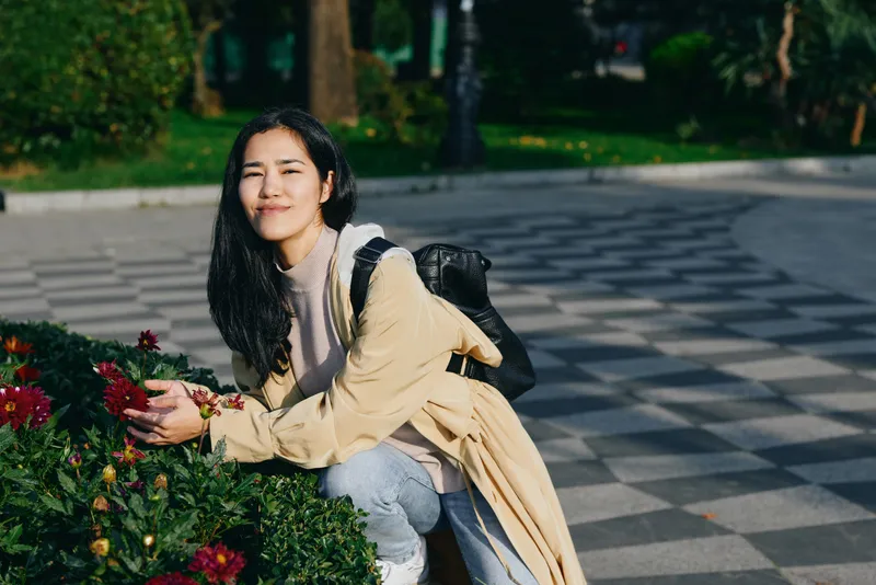 Woman crouching beside flowers in a park, outdoor scene, wearing a beige coat and backpack, with a content smile and greenery around