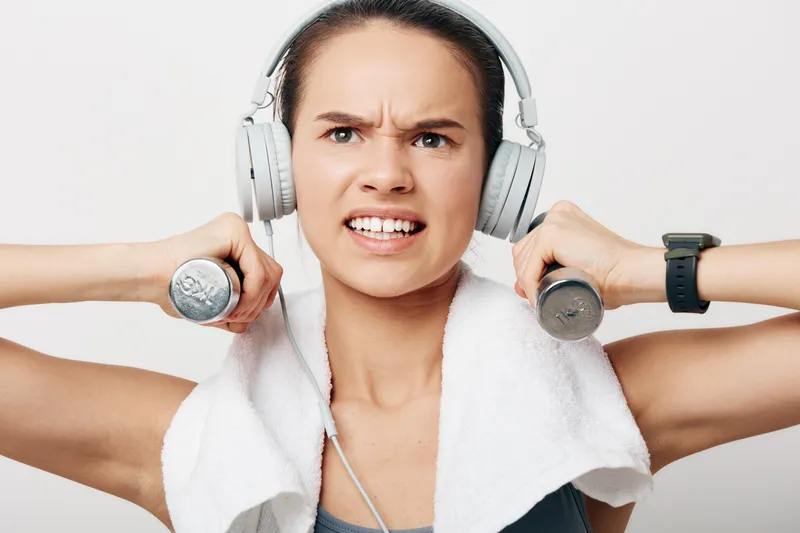 Dynamic portrait of a woman in fitness mode, lifting light dumbbells with a towel around her neck, headphones on, focused gaze, ready for a tough gym workout under bright studio light.