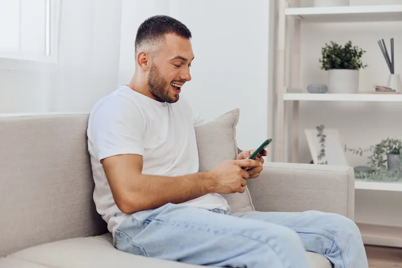Happy man texting on smartphone while sitting on a cozy couch, enjoying quality time at home with modern interior and bright atmosphere.