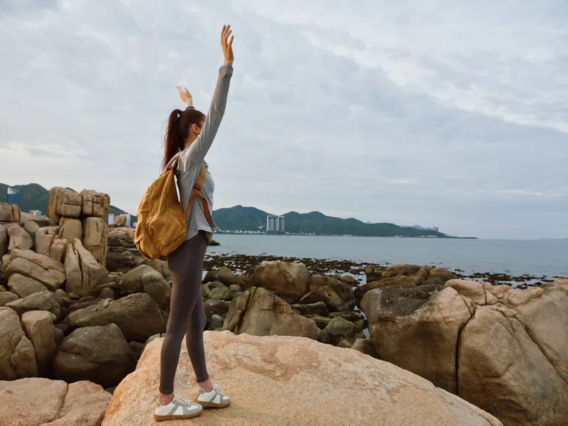 Young woman enjoying nature by the sea, wearing casual outdoor clothing and backpack, expressing joy on rocky shoreline, perfect for travel and adventure themes