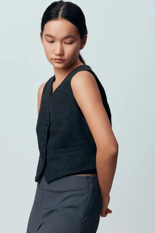 Young woman with black hair wearing sleeveless black vest and gray pants posing on light background. Fashion model looking down with hands behind back, studio portrait of asian female.