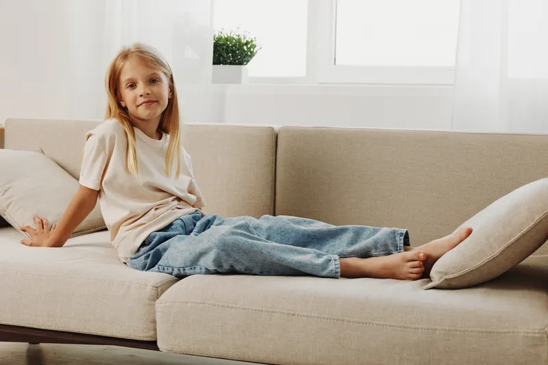 Relaxed young girl with long blonde hair sitting on beige couch in bright living room. Casual outfit and natural light create calm and comfortable home atmosphere.