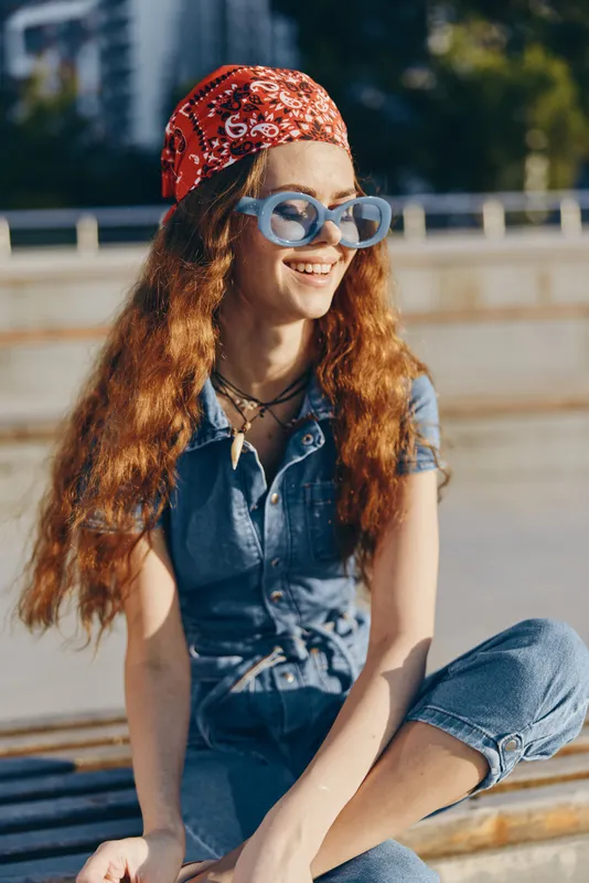 Smiling woman with long red hair wearing sunglasses and a red bandana enjoys a sunny day outdoors, dressed in casual denim outfit with relaxed posture.