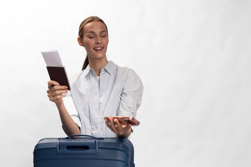 Young woman holding passport and smartphone stands behind blue suitcase with eyes closed, smiling. Concept for travel, vacation, and holiday preparation with positive mood.