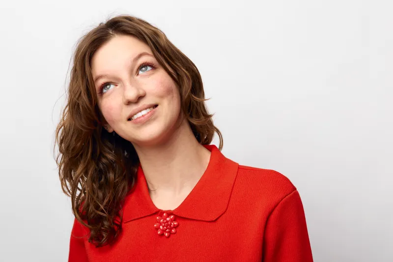 Young woman with curly brown hair wearing red sweater looks up with a smile on white background. Concept of happiness, hope, casual style, and positive emotions in portrait photography.