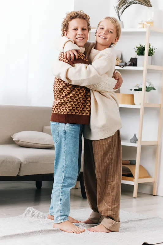 Two smiling children hug warmly in a bright living room, wearing cozy knit sweaters and jeans, conveying affection, comfort, and togetherness in a home setting.