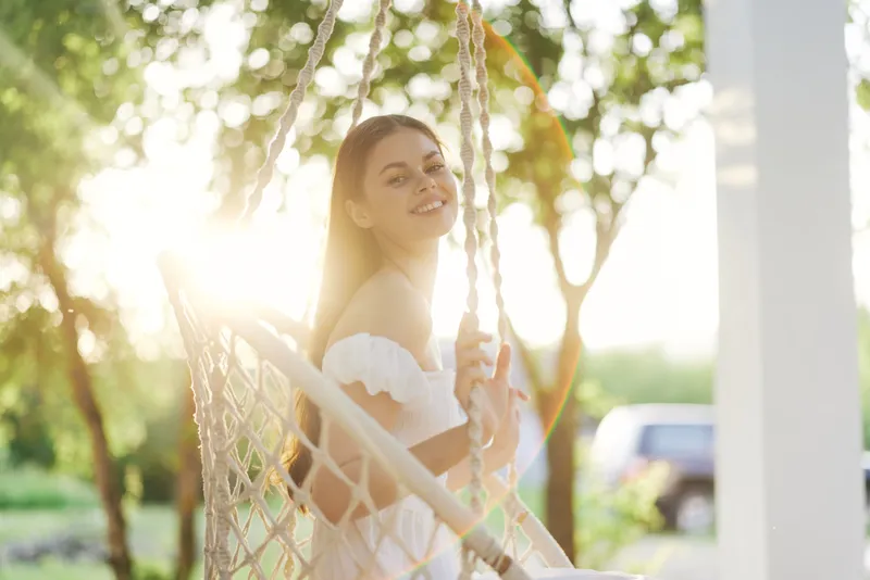 Young woman in a white dress smiling on a swing, surrounded by a bright green garden during sunset, reflecting happiness and tranquility in nature