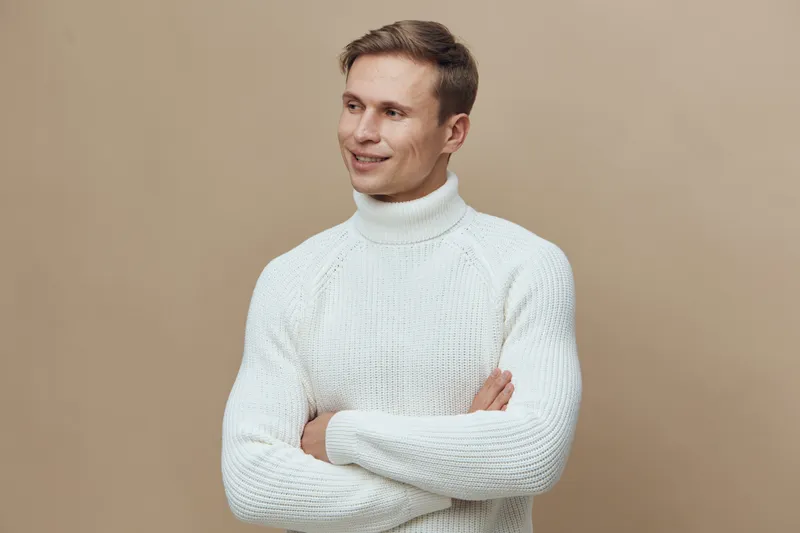 Smiling man wearing white knit sweater standing against a plain beige background. Studio portrait of a cheerful young male with a friendly expression, showcasing casual winter fashion in a bright sett