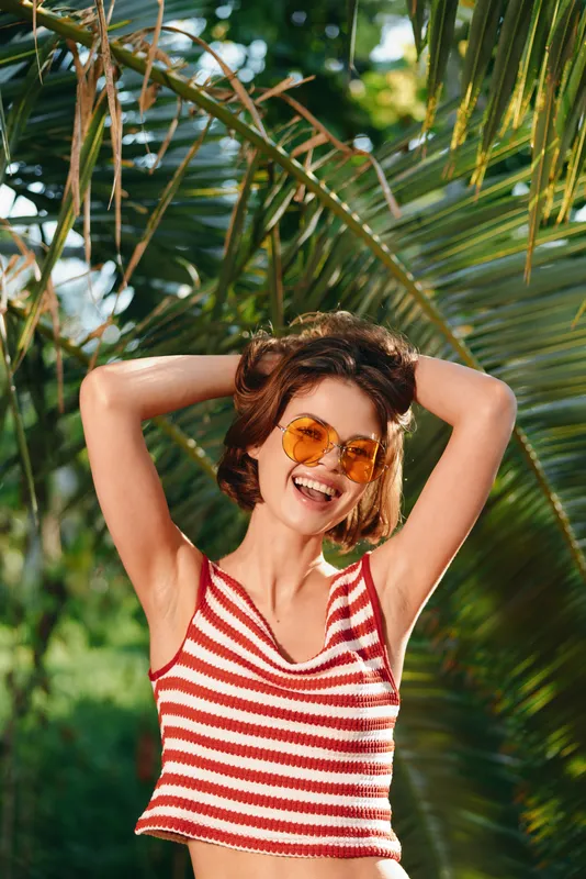 A cheerful woman with striped crop top and sunglasses enjoys a sunny outdoor setting, radiating joy as palm trees frame a vibrant tropical scene.