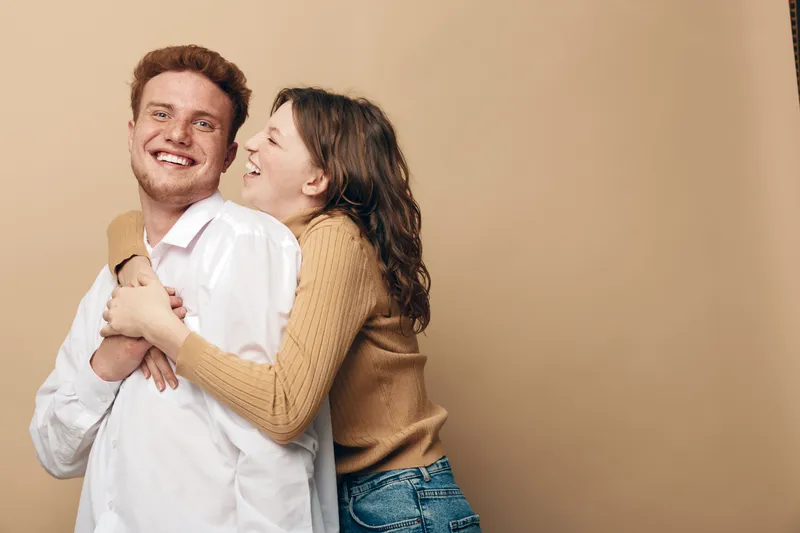 Happy young couple embraces and smiles in casual clothes against beige background. Concept of love, friendship, relationship, and joyful moments together in studio setting.