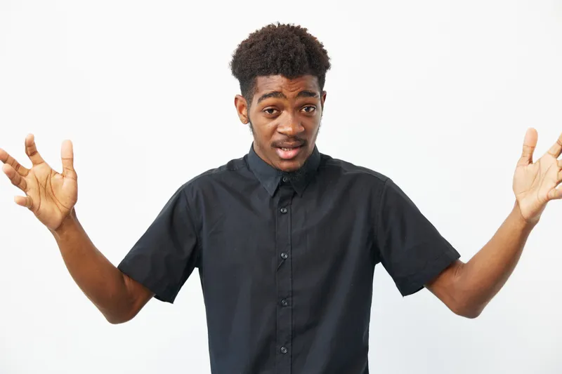 A young African American man gesturing with hands, expressing surprise and enthusiasm. He wears a black shirt against a clean white backdrop, creating a vibrant contrast.