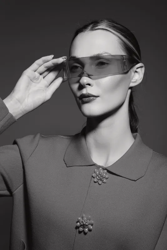 Portrait of a slender Caucasian woman with pale skin wearing futuristic sunglasses and a textured jacket, posing in a studio with dramatic black and white lighting.