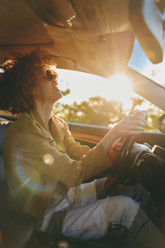 Smiling woman with curly hair driving a car during golden hour. Sunlight fills the cabin with a warm glow, creating a relaxed lifestyle moment focused on travel and self care.