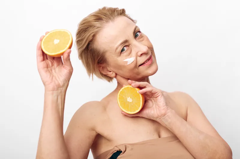 Mature woman with natural skin holds two orange halves near face while applying white cream on cheek. Concept of skincare, healthy lifestyle, natural beauty, and citrus facial treatment.