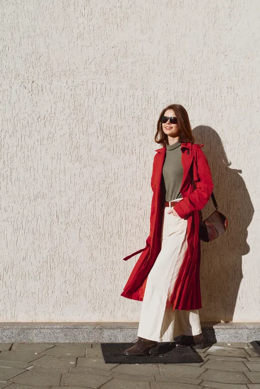 Young woman wearing sunglasses and a red coat walking on a sunny day near a textured wall. Casual outfit with white pants and brown shoes. Urban lifestyle concept.