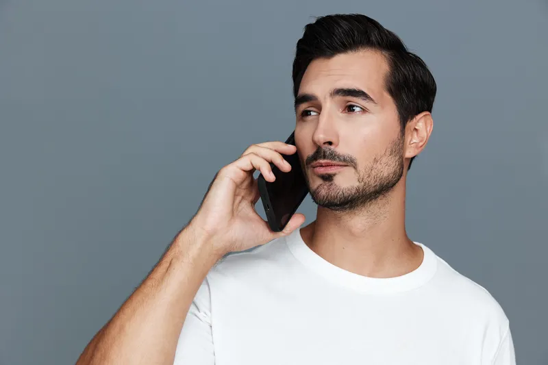 Thoughtful young man wearing casual white t-shirt using smartphone while looking away against a plain grey background. Modern lifestyle concept.