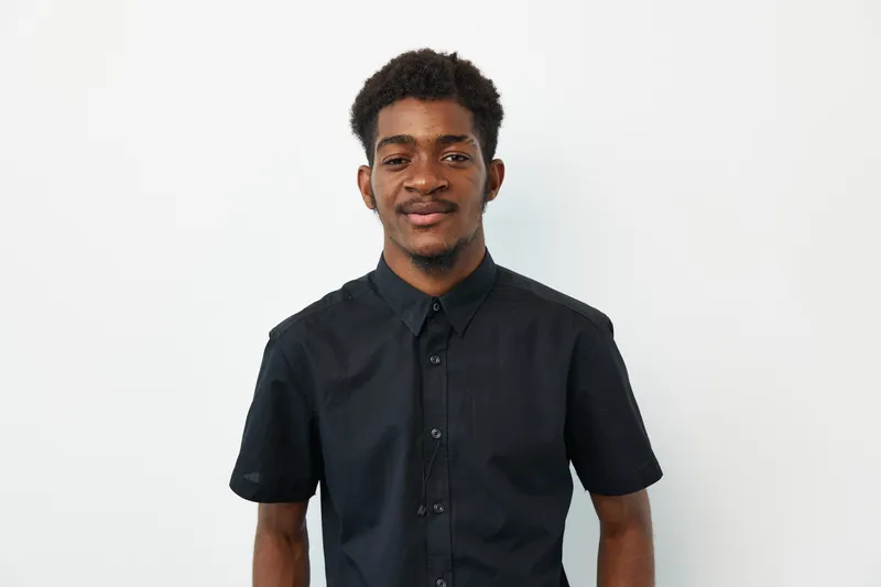 Confident black man with a stylish haircut wearing a black shirt. His expression exudes positivity and enthusiasm, set against a clean white backdrop.
