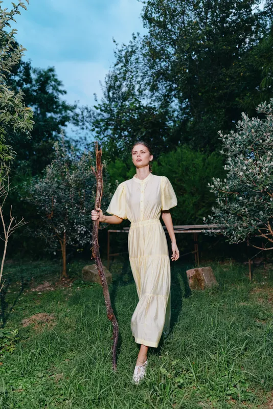 A woman in a pale dress walks through a grassy garden holding a long stick, surrounded by trees and a clear sky, creating a calm natural fashion moment outdoors with soft light