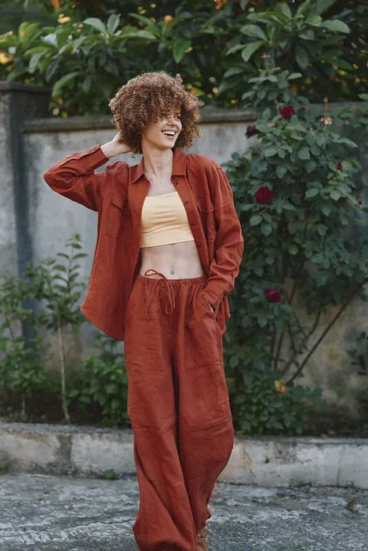 Smiling woman with curly hair wearing a rust colored suit outdoors, posing for a fashion lifestyle shot. Natural garden backdrop, relaxed confidence, casual chic, warm daylight