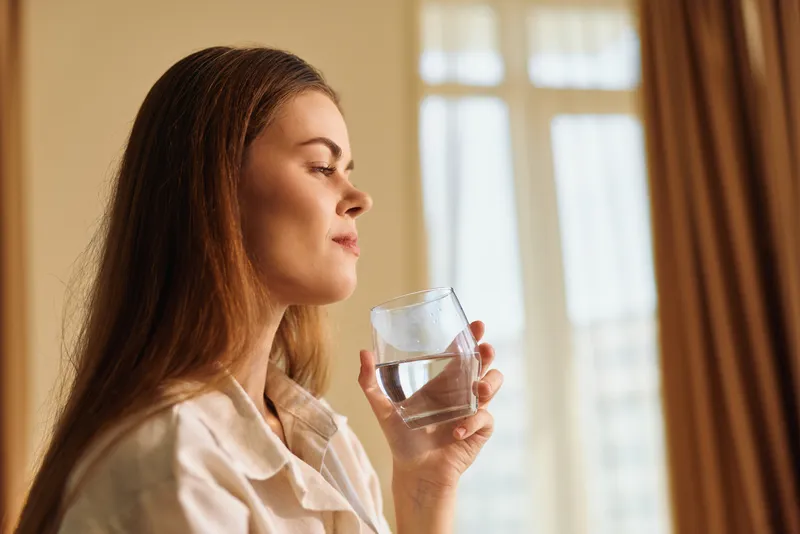 woman glass water drink portrait home relaxation morning - Closeup profile of a woman in pajamas holding a clear glass of water by a window, sipping for hydration and peaceful morning routine.