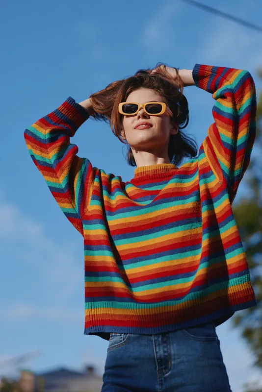 woman, rainbow sweater, sunglasses, outdoor, smiling, casual outfit, young adult, blue sky — Young adult woman in a colorful striped sweater and yellow sunglasses posing outdoors, smiling confidently