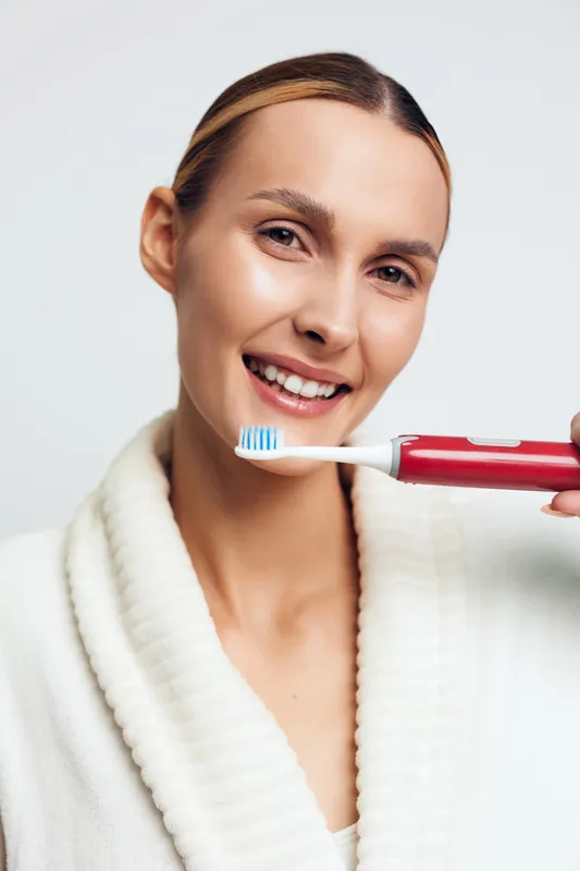 A smiling young woman in a white bathrobe holds a red electric toothbrush near her mouth. The close-up portrait shows dental hygiene and morning routine concepts with clean skin and natural light.