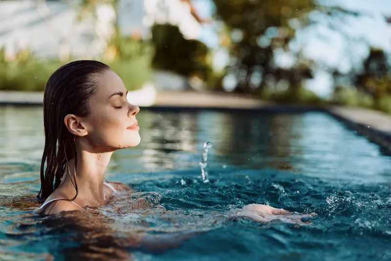 Relaxed pool scene shows a woman with closed eyes enjoying calm water and sunshine, a serene moment of relaxation, leisure, and wellness beside a tranquil outdoor pool in a private lounge area